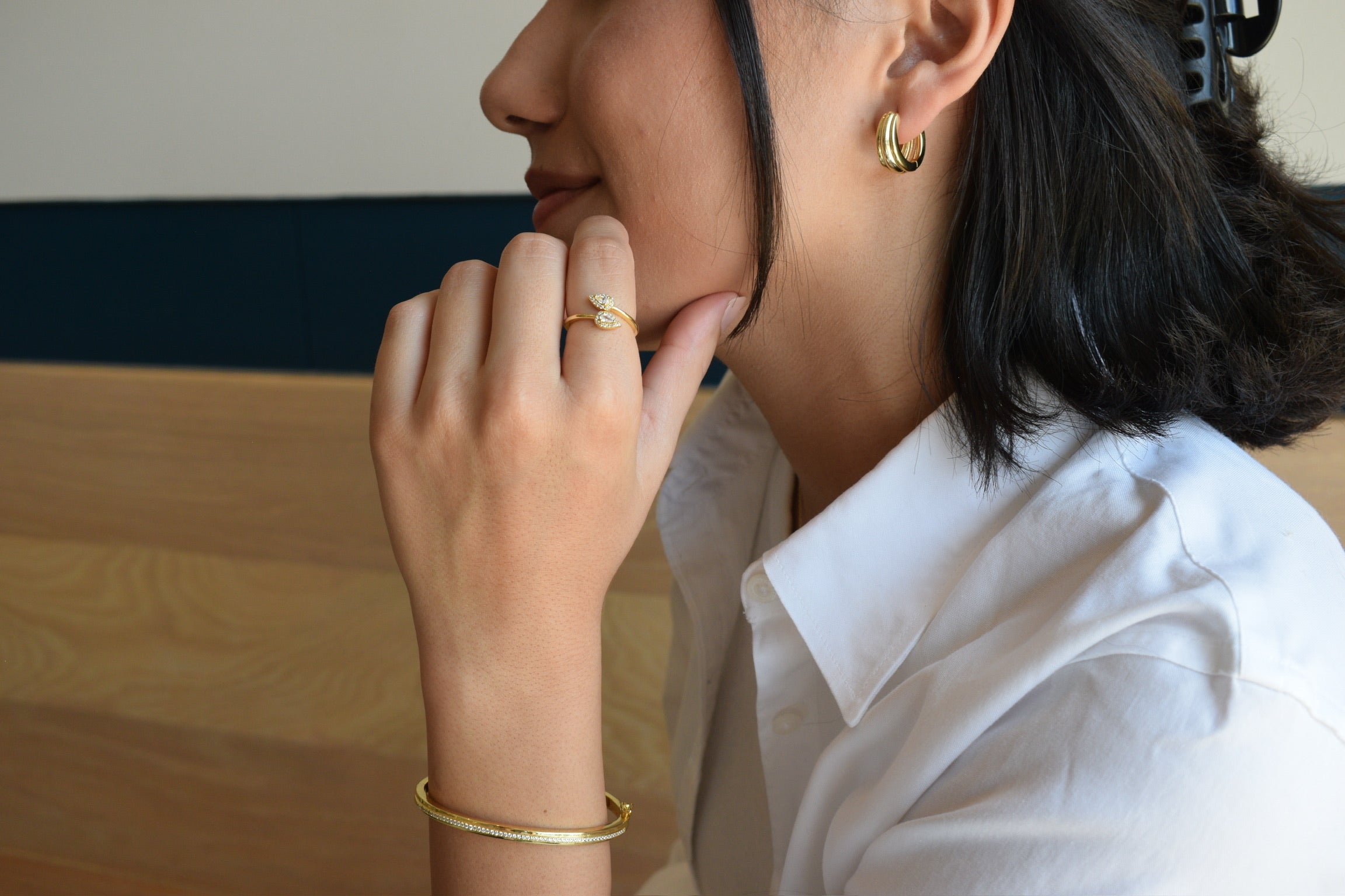 A person wearing a gold-colored bangle on their left wrist, with an oval shape and a sleek design.